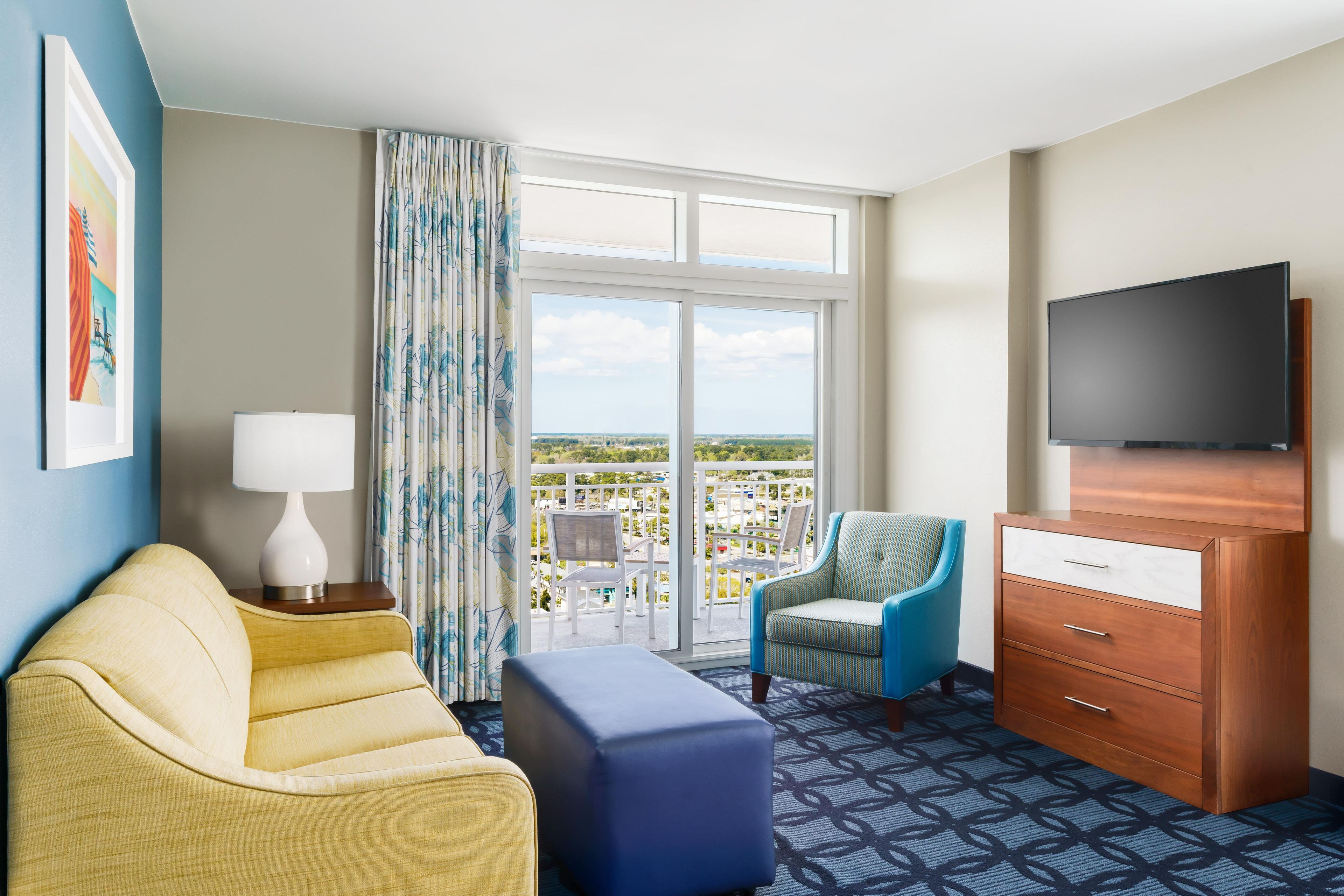 Een hotelkamer met een bank, een stoel, een televisie, een dressoir en een balkon met uitzicht.