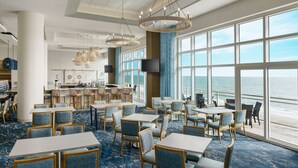 A spacious dining area with tables and chairs, large windows with a view of the ocean, and a bar area in the background.