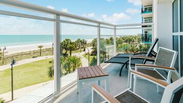 A balcony with a view of the beach, a table, and two chairs.