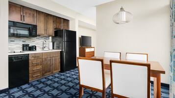 A modern kitchen with wooden cabinets, a black refrigerator, and a microwave. There is a dining table with chairs and a hanging light fixture.