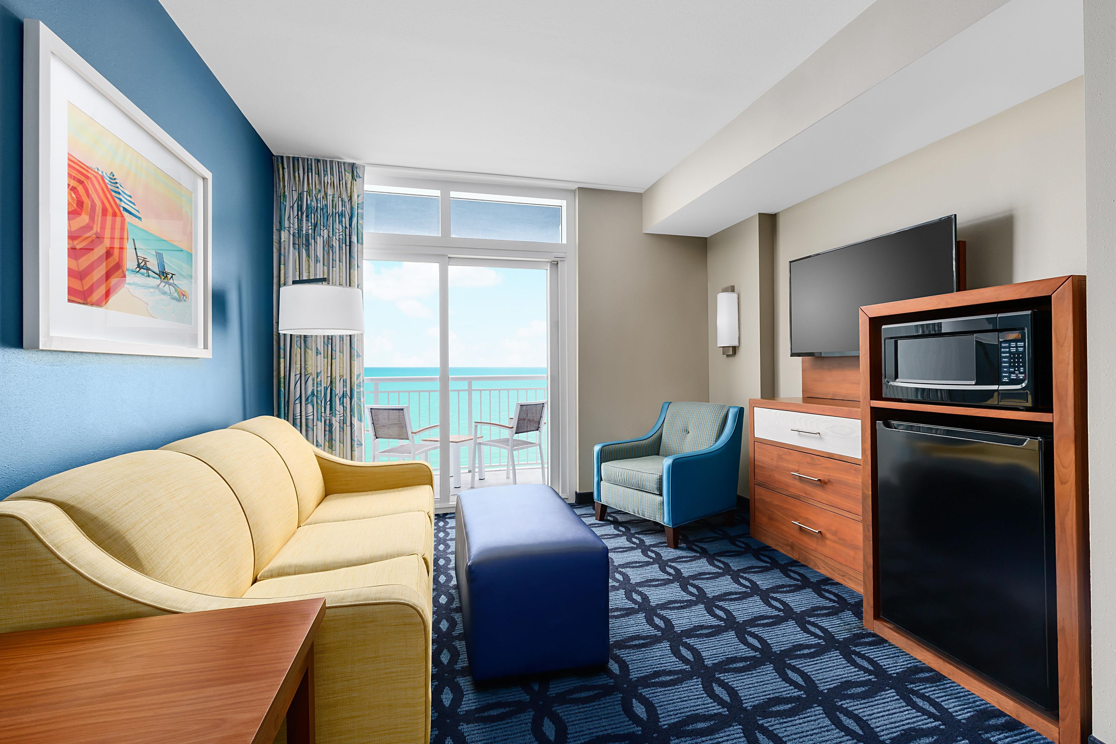 A hotel room with a sofa, a chair, a desk, a television, a microwave, and a view of the ocean.