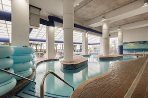 An indoor swimming pool with multiple lanes, a lounge area with chairs, and a view of palm trees through large windows.