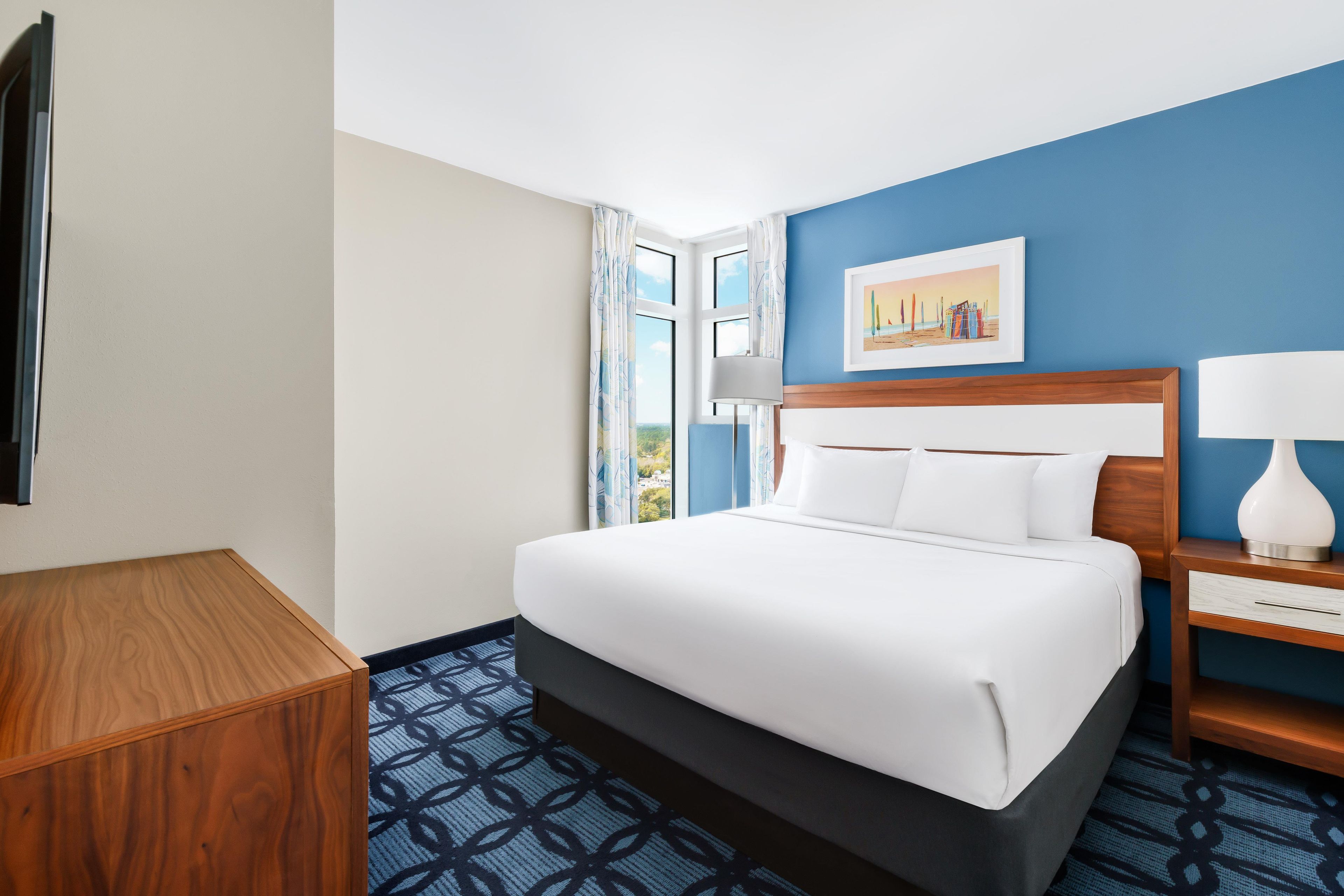 A hotel room with a bed, a nightstand, a lamp, a painting on the wall, and a window with curtains.