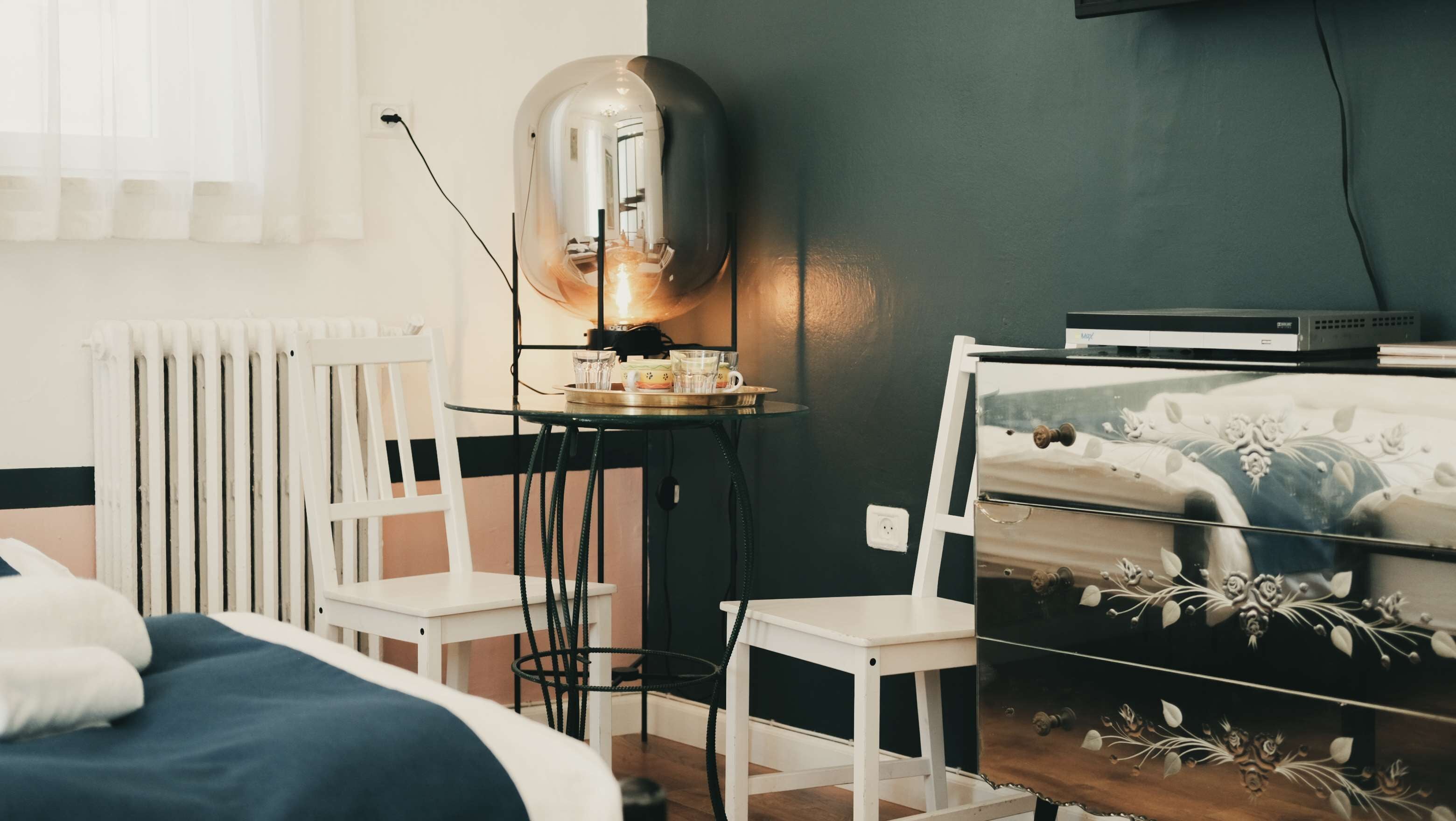 A bedroom with a bed, a radiator, a small table with cups and a kettle, a chair, and a dresser with decorative items.