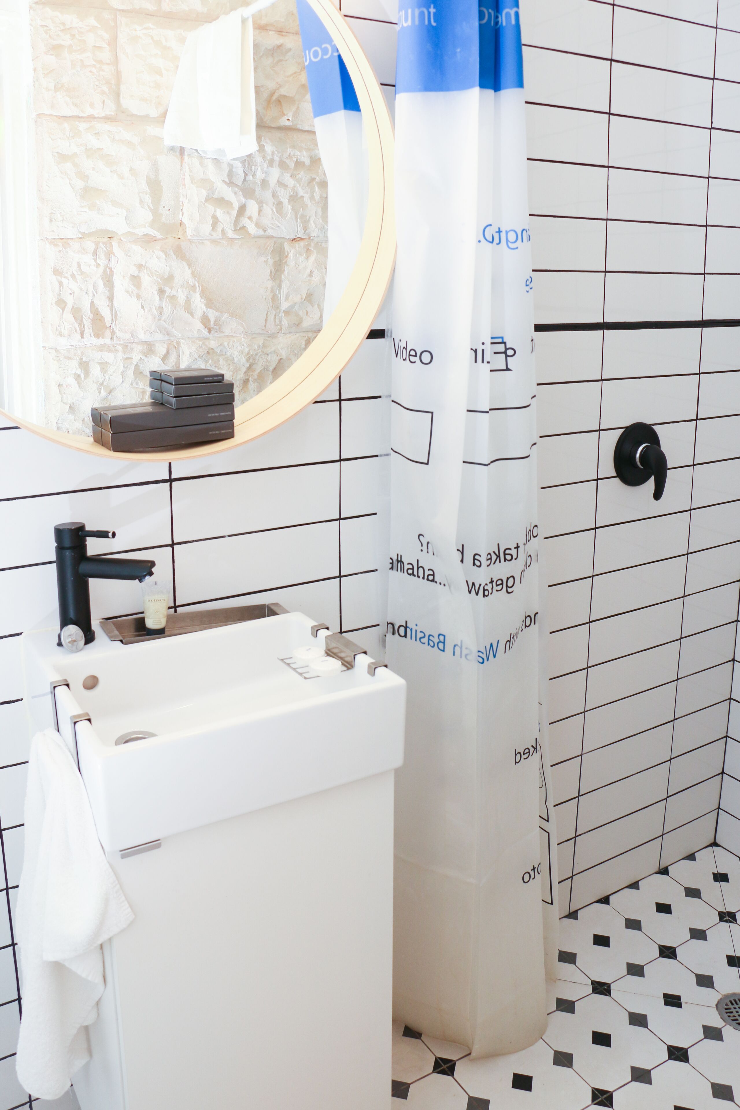 Une salle de bain avec un lavabo blanc, un robinet noir et un miroir au-dessus. Il y a un rideau de douche avec du texte imprimé dessus, et le sol est recouvert d’un motif à carreaux noirs et blancs.
