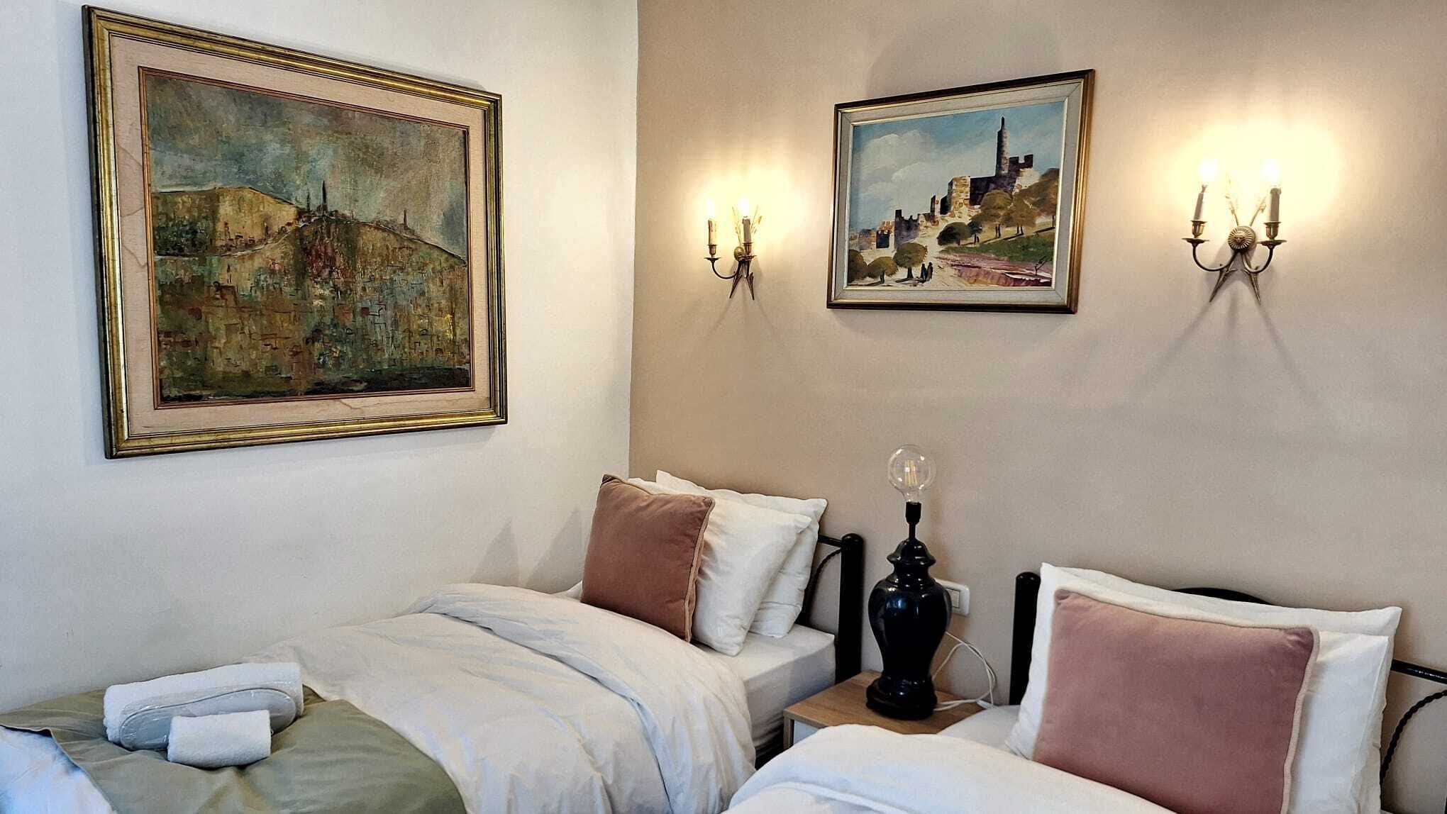 A hotel room with two beds, a painting on the wall, and two wall-mounted lamps.