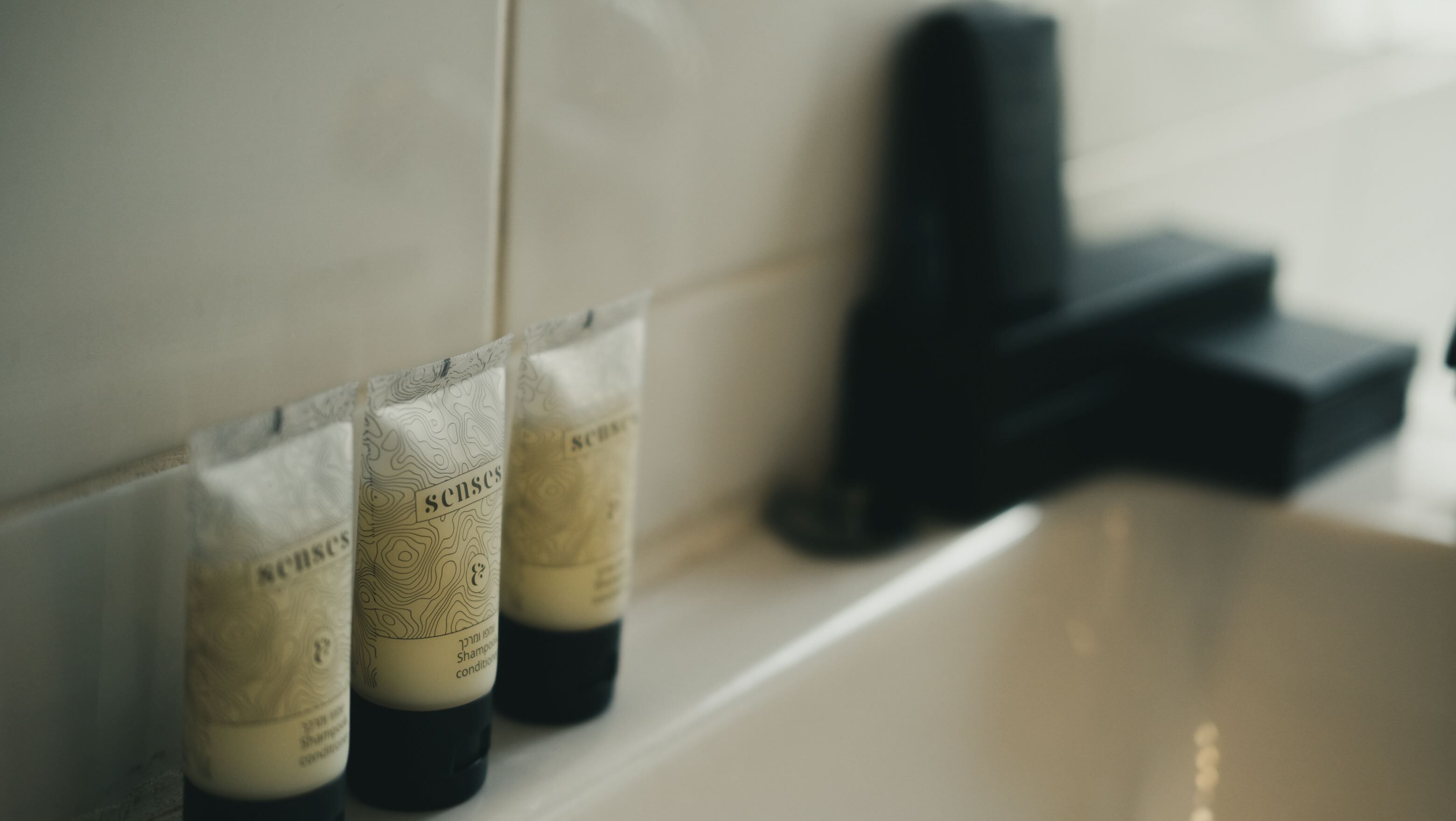 Un baño con un lavamanos blanco, un grifo negro y tres botellas de champú con la etiqueta 'senses' en la pared.