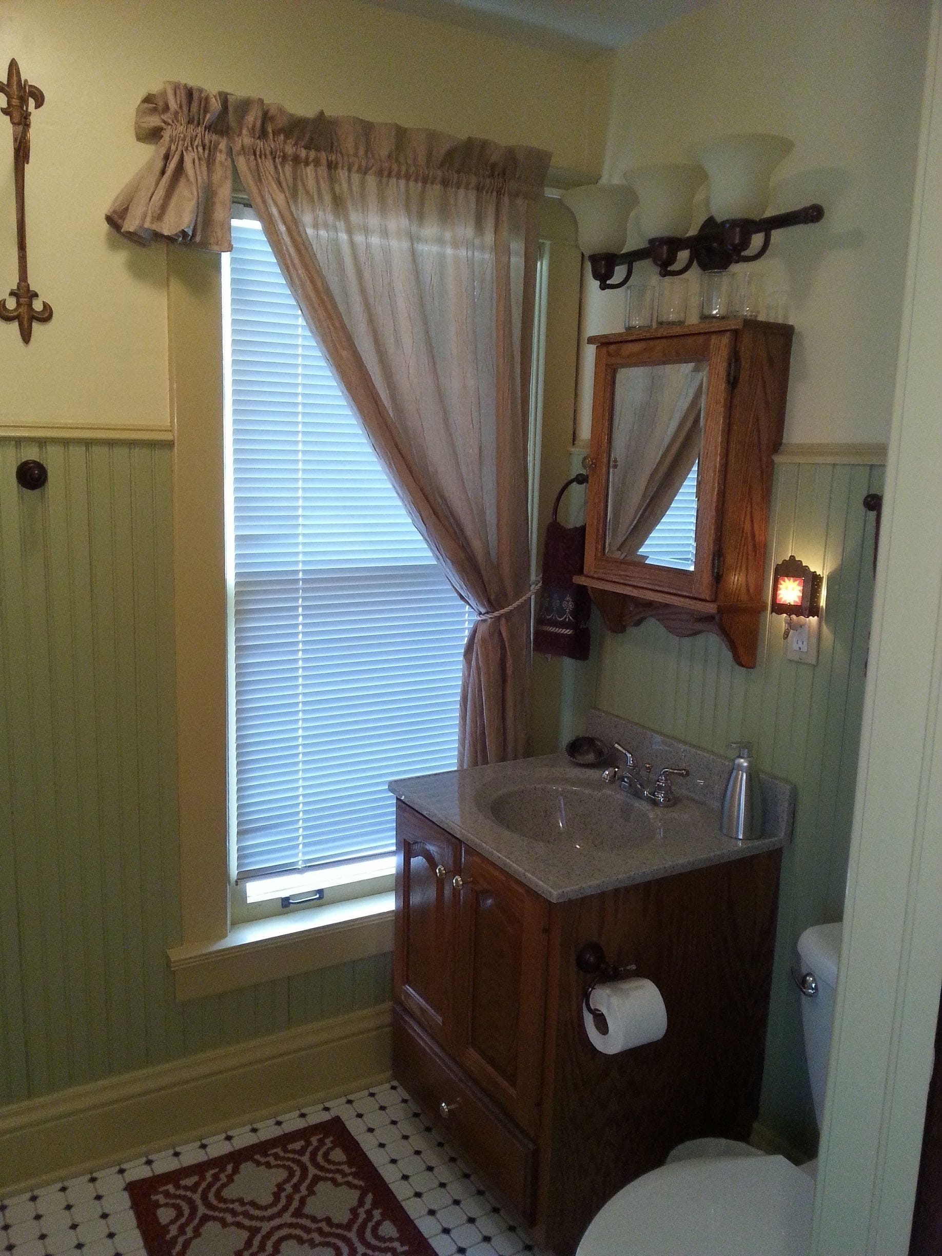 A bathroom with a vanity, sink, mirror, toilet, and window with curtains.