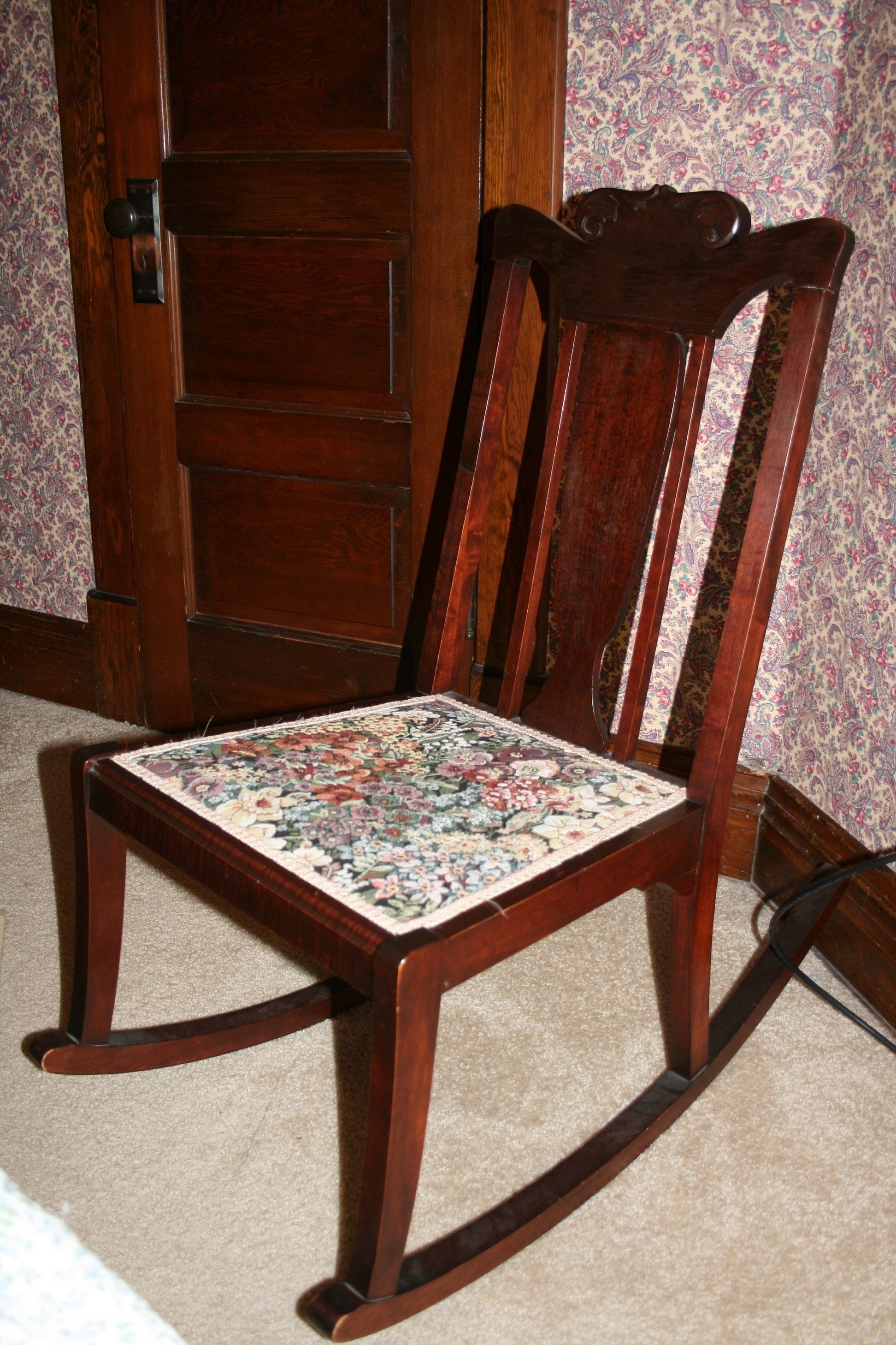 Een houten schommelstoel met een kussen met bloemenmotief, geplaatst in een kamer met behang met een patroon en een houten deur.