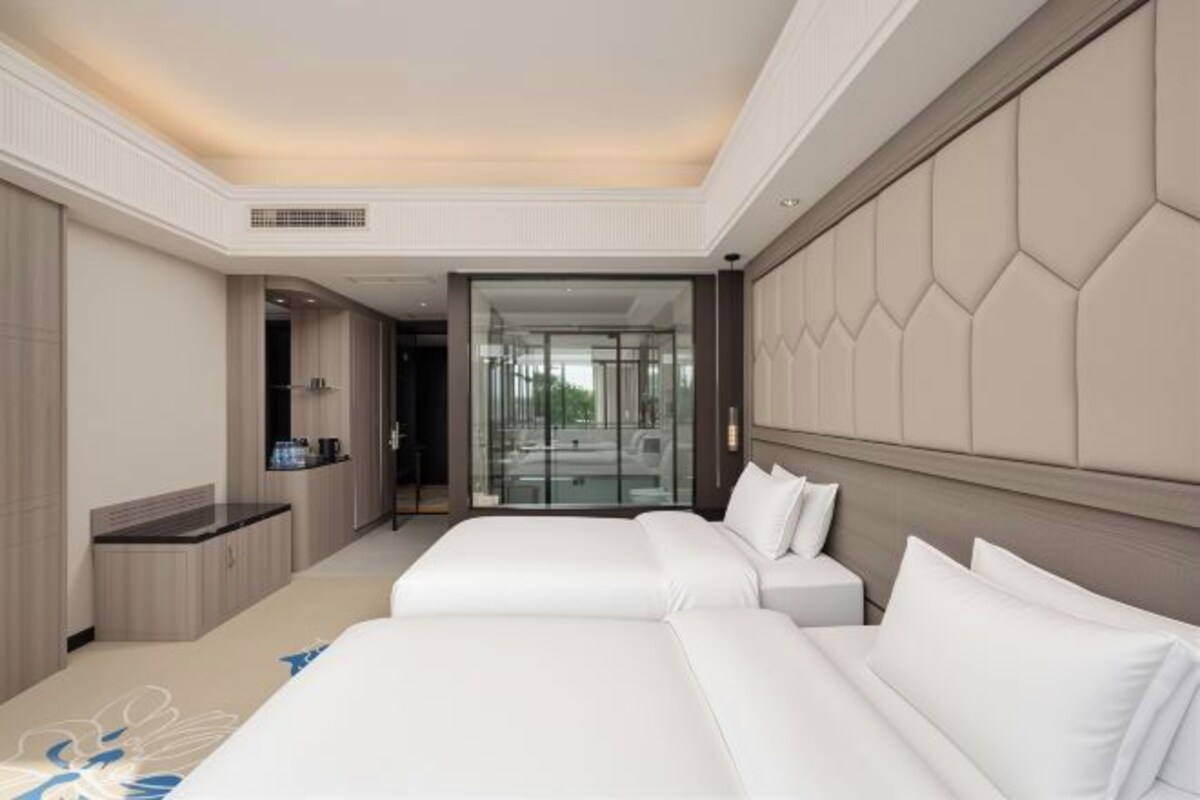 A hotel room with two beds, a sitting area, a glass-enclosed bathroom, and a view of the outdoors.