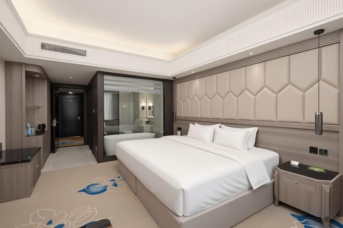 A modern hotel room with a large bed, bedside tables, a built-in wardrobe, and a glass-enclosed bathroom.
