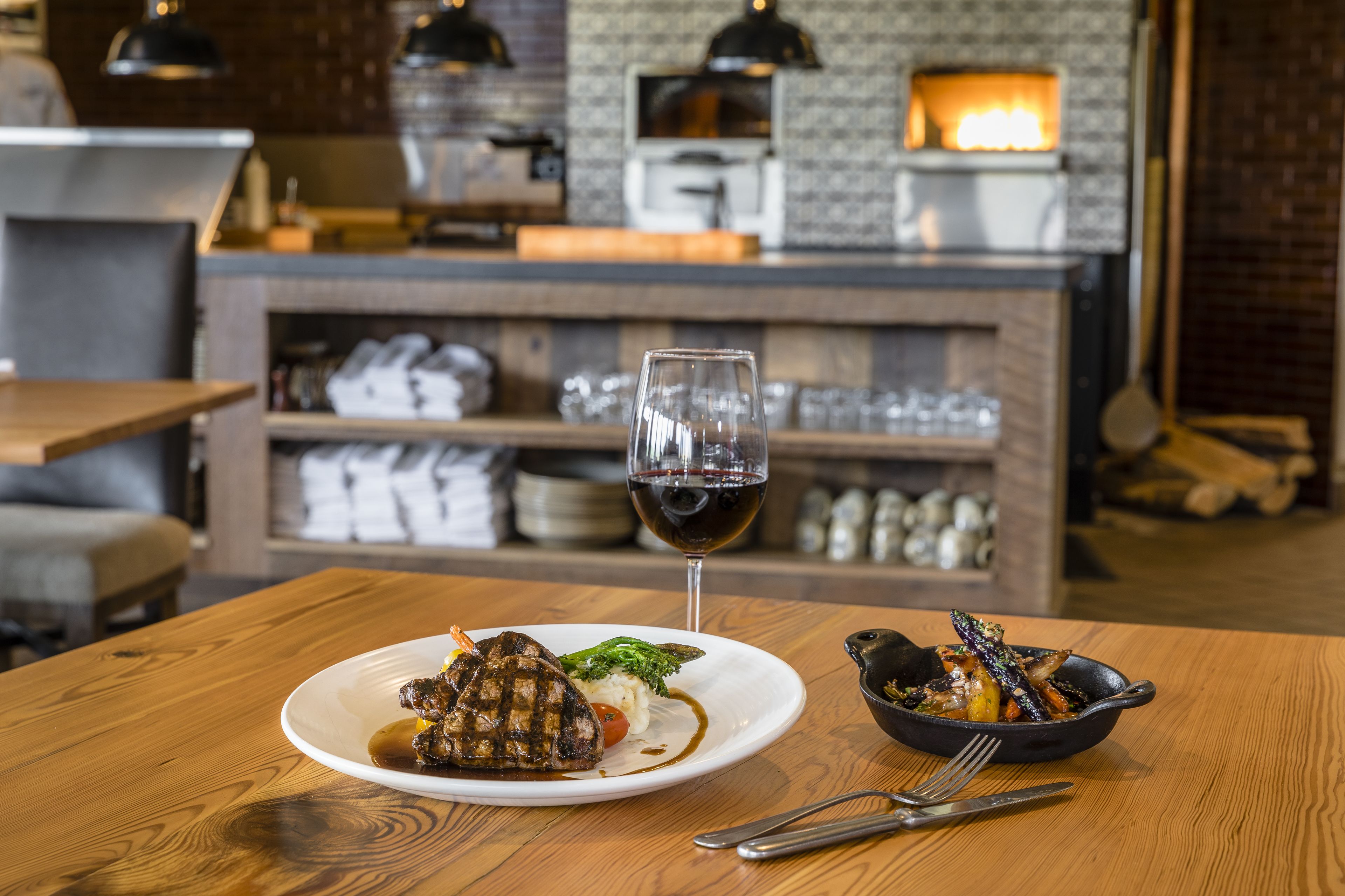 A restaurant setting with a wooden table, a plate of food, a glass of red wine, and a bowl of vegetables.