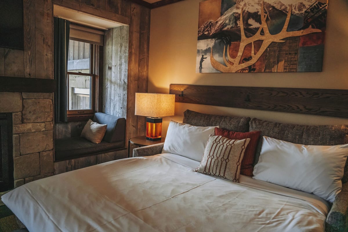A bedroom with a large bed, a nightstand with a lamp, a window with a view, and a painting on the wall.