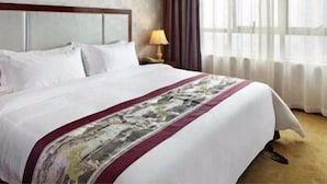 A hotel room with a bed, a painting on the wall, a window with curtains, and a lamp on the nightstand.