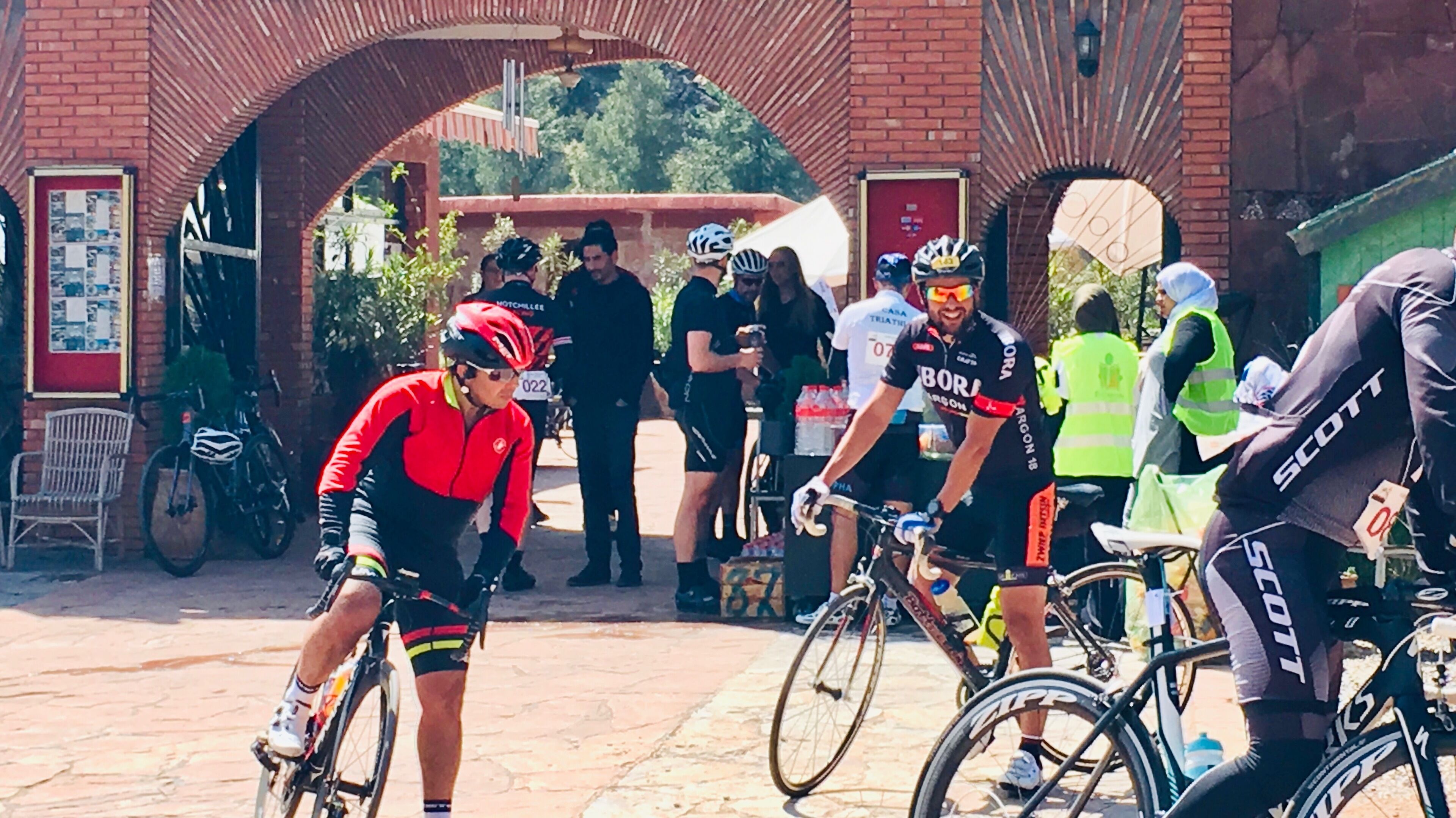 A group of cyclists at a starting line with a brick archway and a signboard in the background.