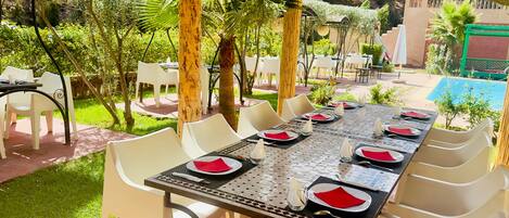 Un área de comedor al aire libre con una mesa larga dispuesta para seis personas, rodeada de sillas blancas.