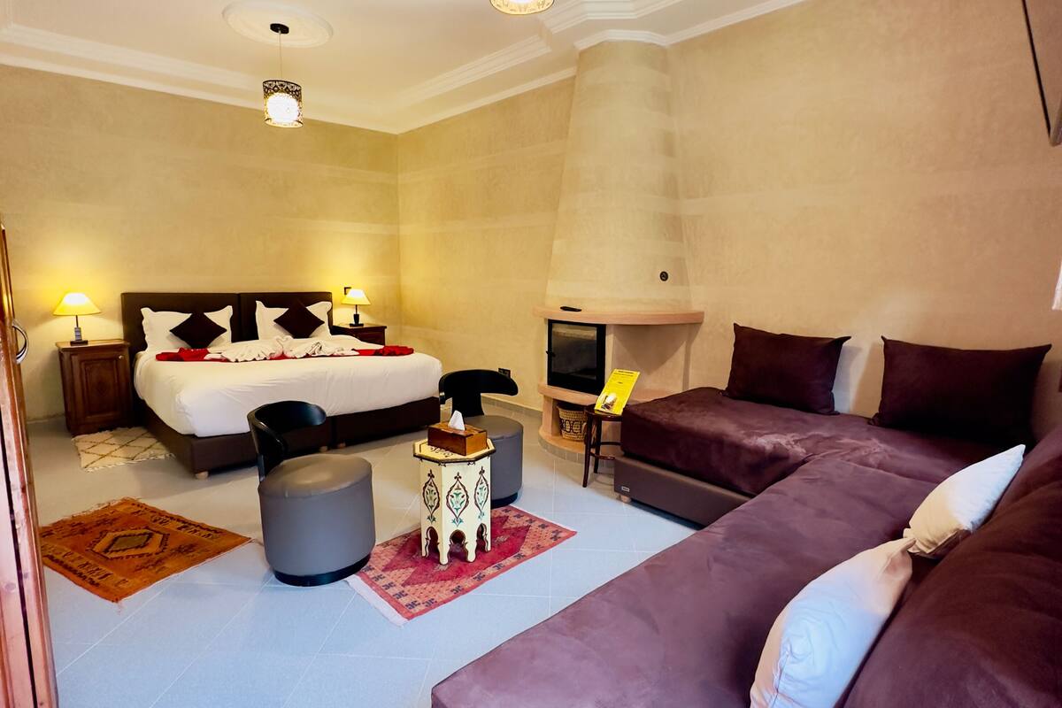 A hotel room with a large bed, a sofa, ottomans, a fireplace, and a television.
