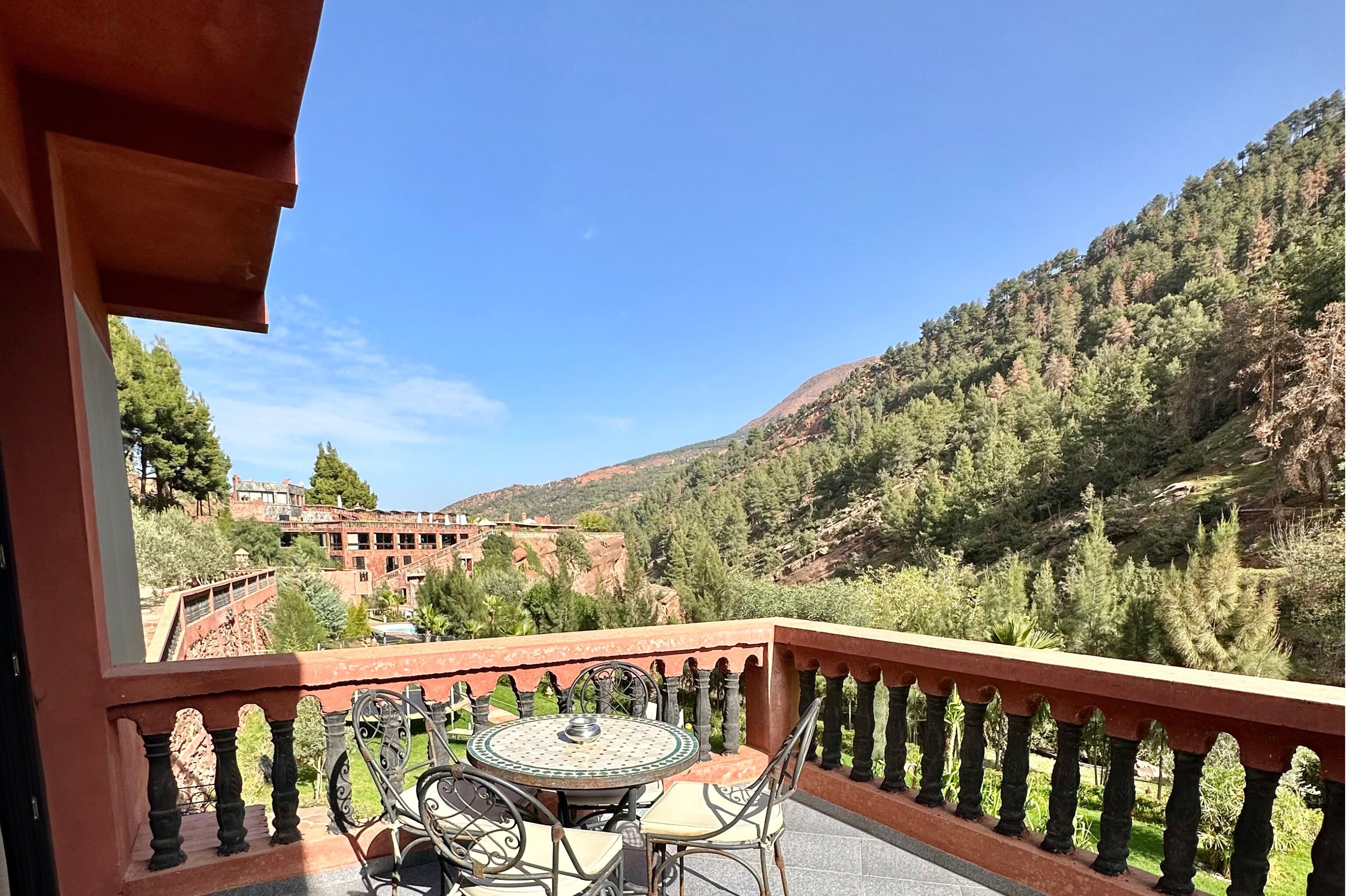A balcony with a table and chairs, overlooking a mountainous landscape with buildings in the distance.