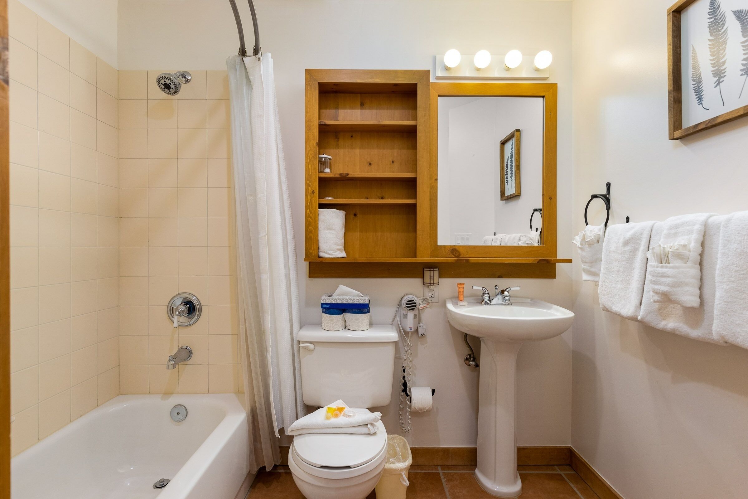 Une salle de bain avec une baignoire, des toilettes, un lavabo et une étagère en bois au-dessus du miroir.