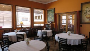 Interior de restaurante con varias mesas redondas, sillas, ventilador de techo y ventanas grandes con persianas.