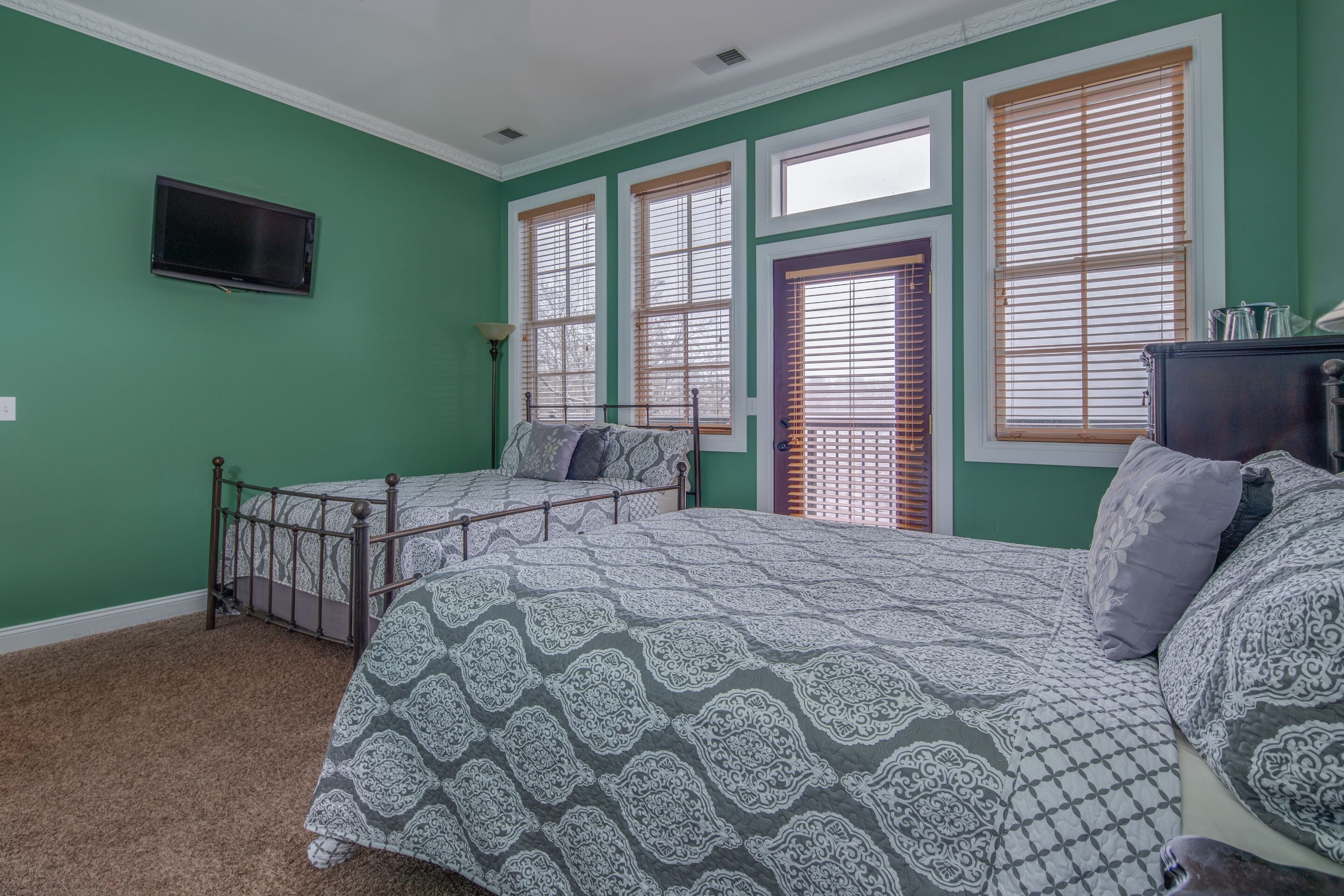 A bedroom with two beds, green walls, and large windows with blinds.