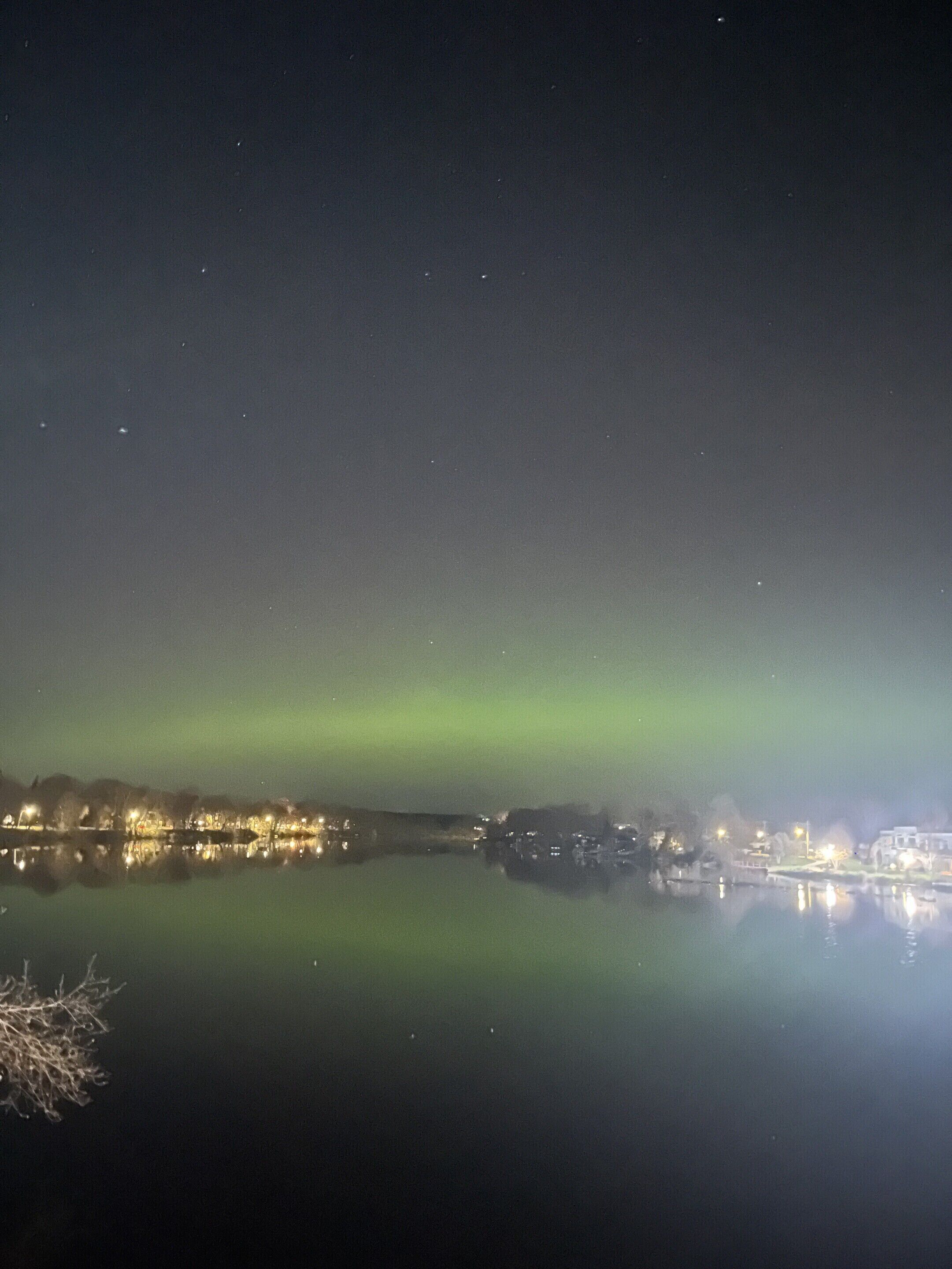 A nighttime view of a waterfront with buildings reflecting on calm water, a visible aurora, and a clear sky.