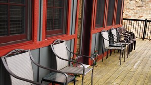 Una hilera de sillas metálicas con asientos de mimbre sobre una terraza de madera, junto a un edificio con ventanas de marco rojo.