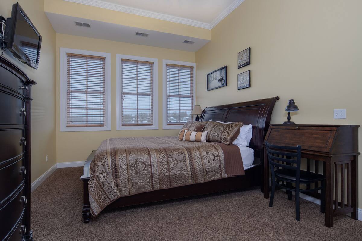 A bedroom with a bed, dresser, desk, and chair. There are three windows with blinds, a lamp, and a television on the dresser.