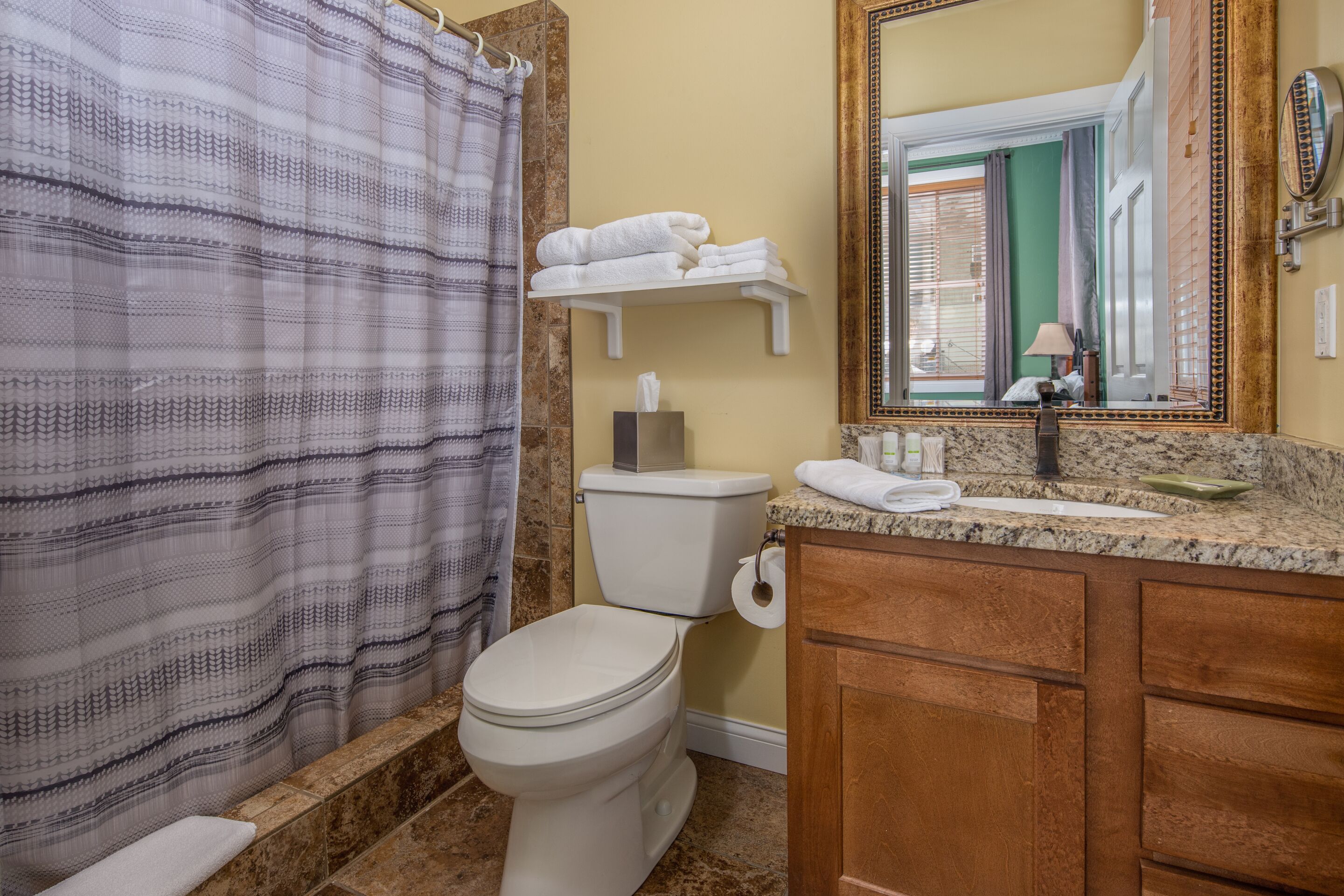 A bathroom with a toilet, a shower curtain, a towel rack with folded towels, a mirror, and a countertop with a soap dish.