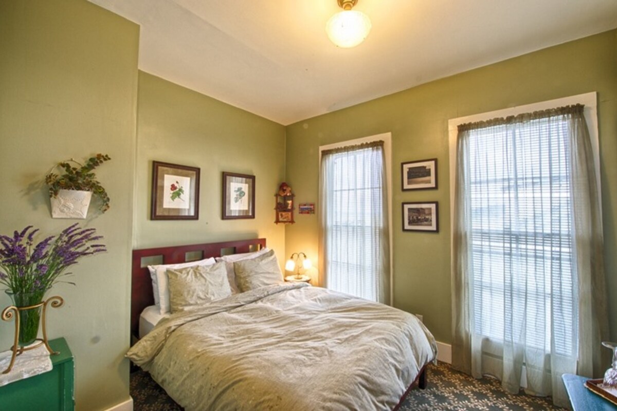 A bedroom with a bed, two framed pictures on the wall, a nightstand with a lamp, and a window with curtains.