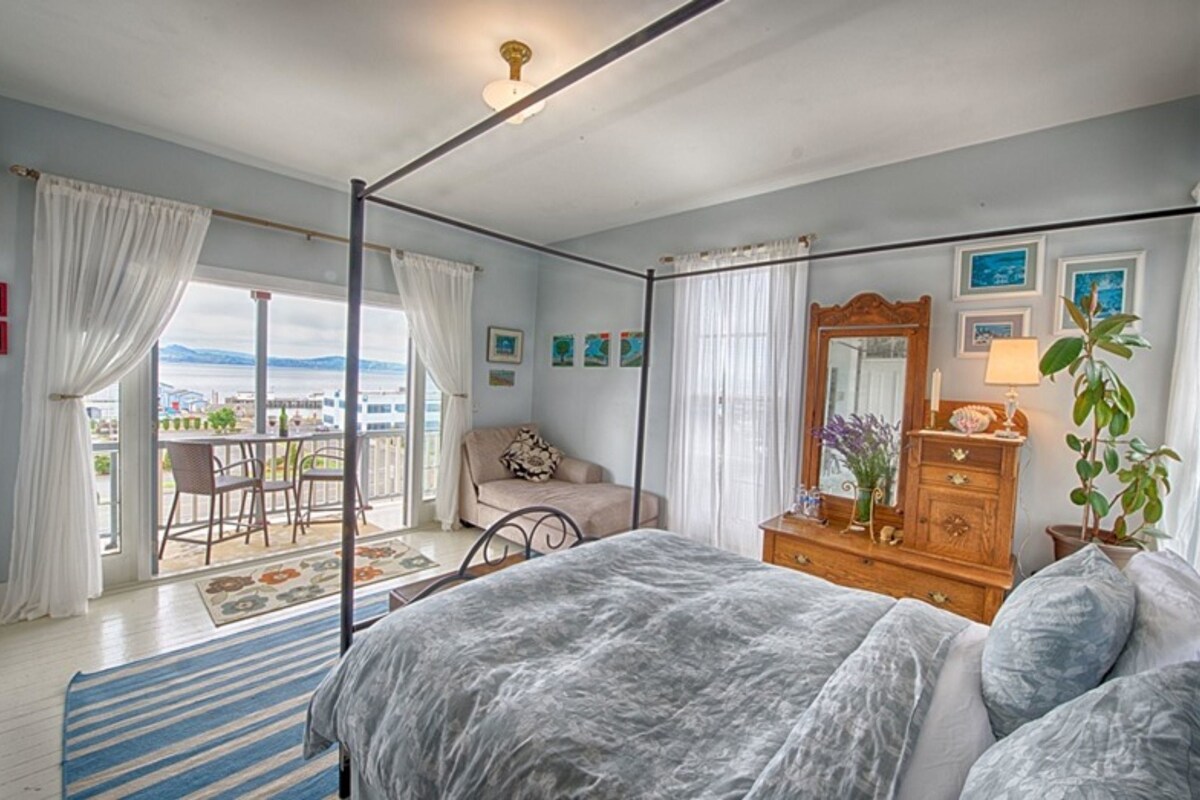 A bedroom with a four-poster bed, a wooden dresser, a mirror, and a view of the outdoors through large windows.