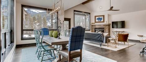 A spacious living room with a large dining table, comfortable seating, a fireplace, and a television mounted on the wall.