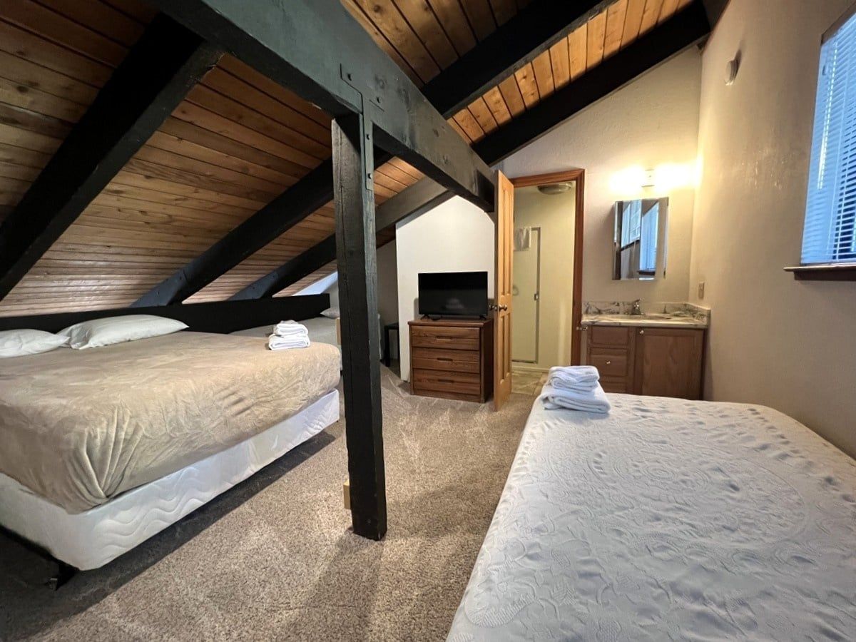 A bedroom with two beds, a wooden ceiling, a small TV, a wooden dresser, and a bathroom with a mirror.