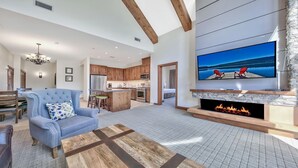 A spacious living room with a fireplace, a television mounted on the wall, and a kitchen in the background.