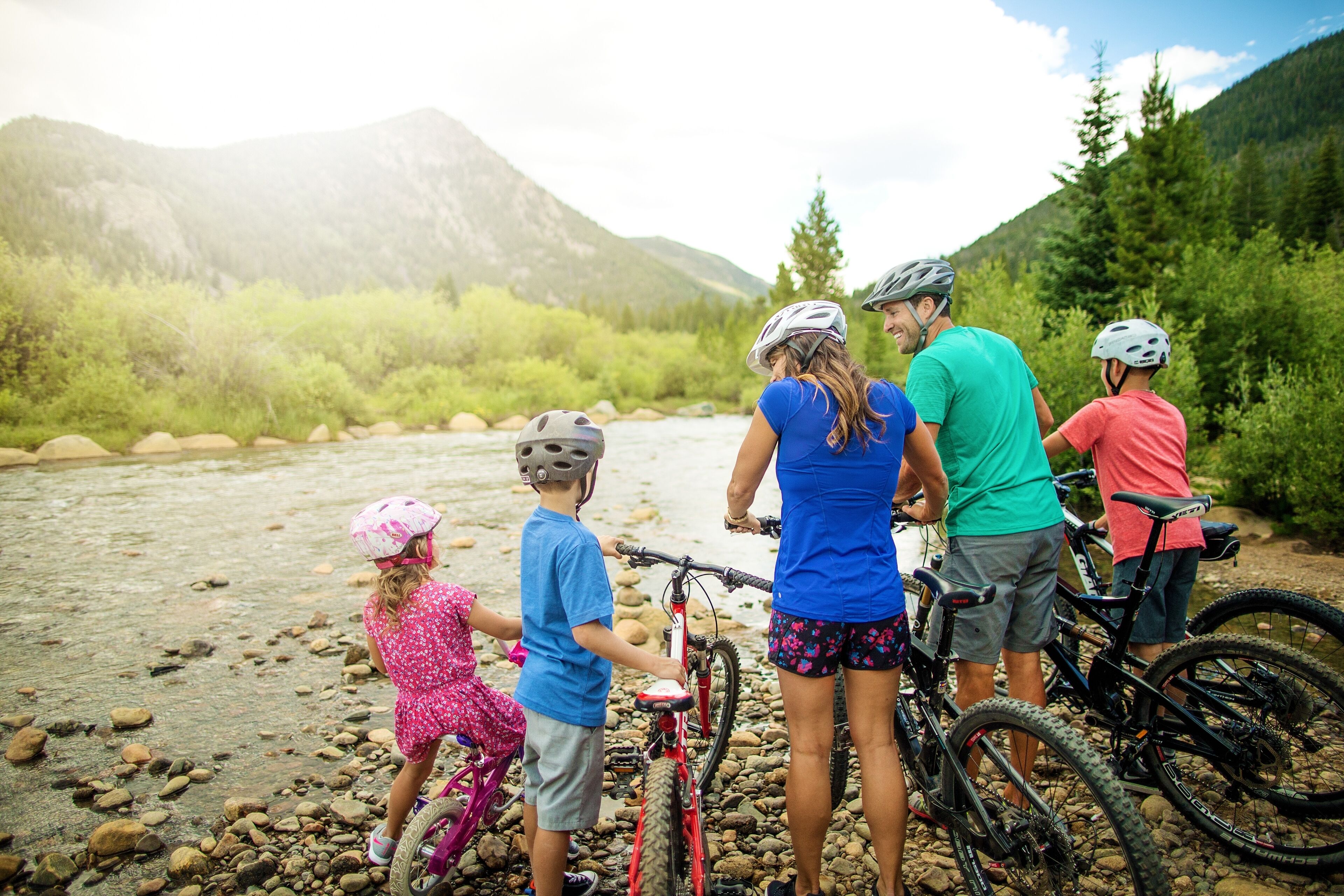 A group of people with bicycles by a river with mountains in the background.