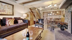A cozy living room with a leather sofa, a wooden coffee table, and a chandelier. A staircase leads to a kitchen with wooden cabinets and a stone wall. - Modern 2 BR Condo Lodge at Vail, Walk to Gondola by RedAwning (Vail)