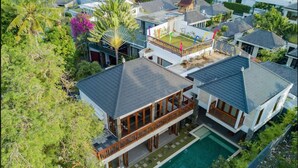 Uma casa de dois andares com piscina, cercada por vegetação exuberante e outros edifícios residenciais.