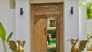Uma porta de madeira com entalhes elaborados, ladeada por duas estátuas douradas de dragão em pedestais, dando acesso a um pátio com uma estátua e uma piscina.