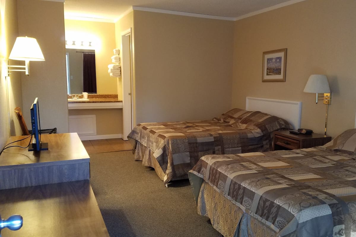 Habitación de hotel con dos camas, un escritorio, una silla, un espejo, un baño y un cuadro en la pared.