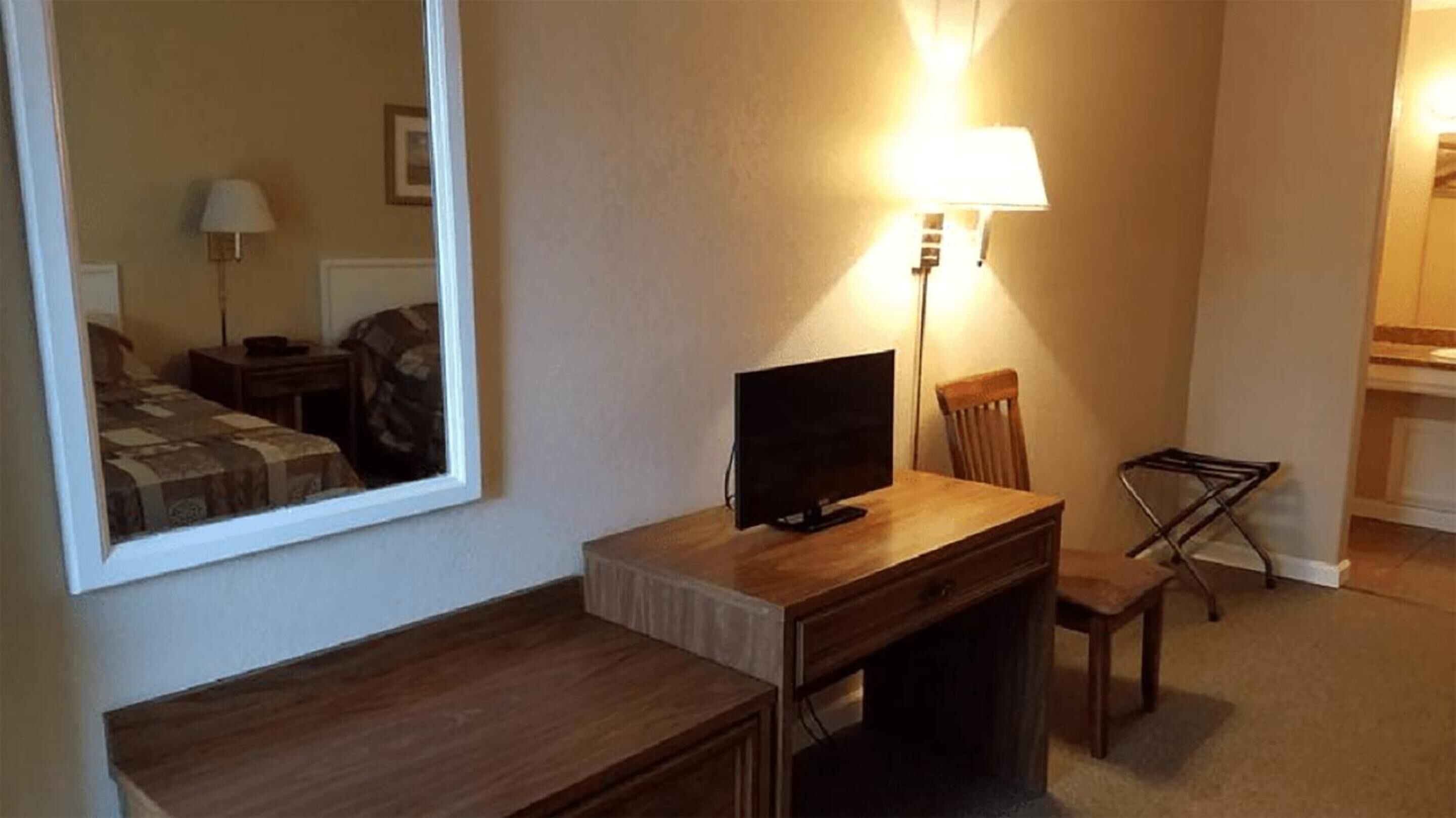 Et hotelværelse med skrivebord i træ, fjernsyn, stol, lampe og spejl.