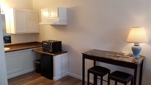 A compact kitchen with a built-in oven, a small table with two stools, a trash bin, and a lamp on a side table.