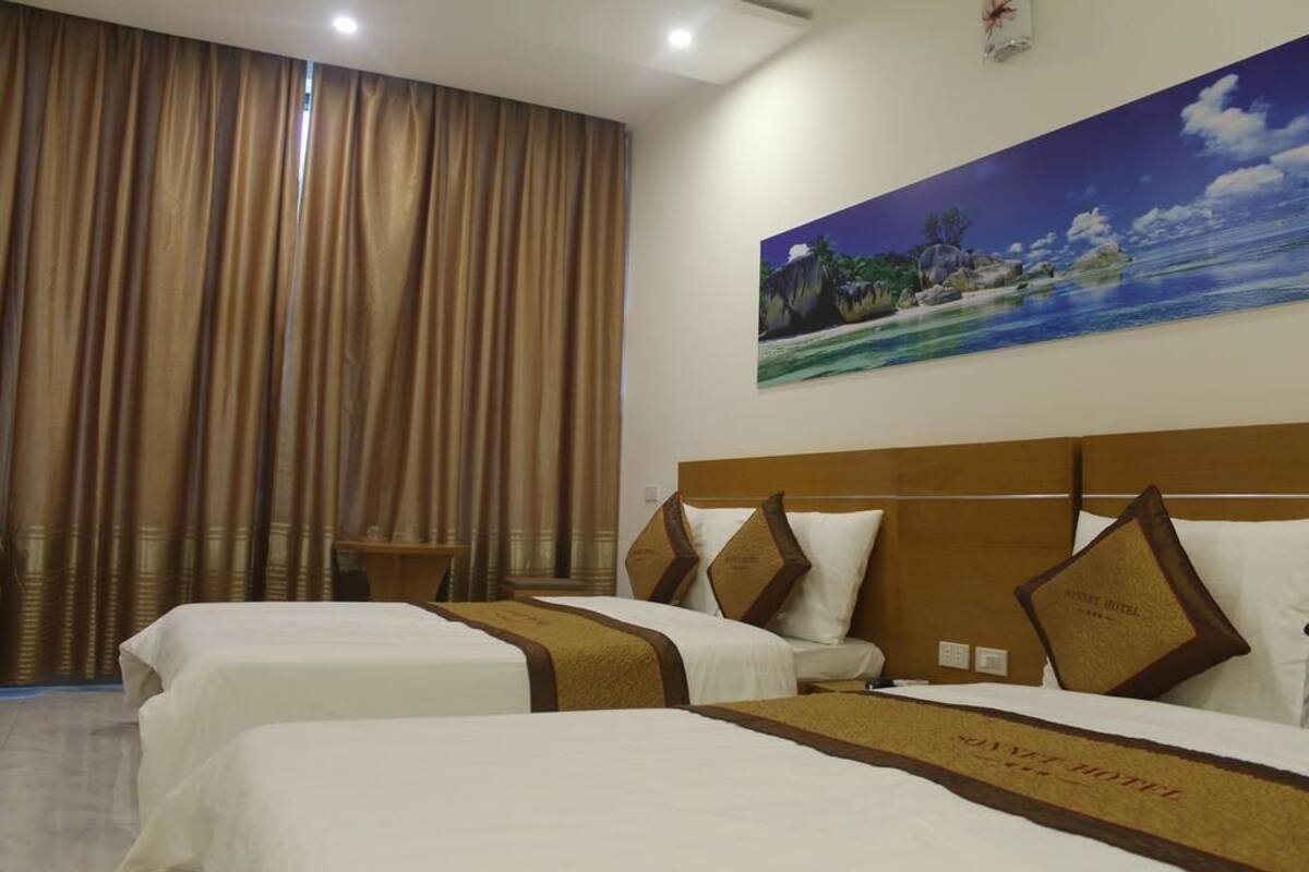 Ein Hotelzimmer mit zwei Betten, einem großen Fenster mit Vorhängen, einem Bild einer tropischen Strandlandschaft und einem Kopfteil mit einem Kopfkissen, das den Namen des Hotels zeigt.