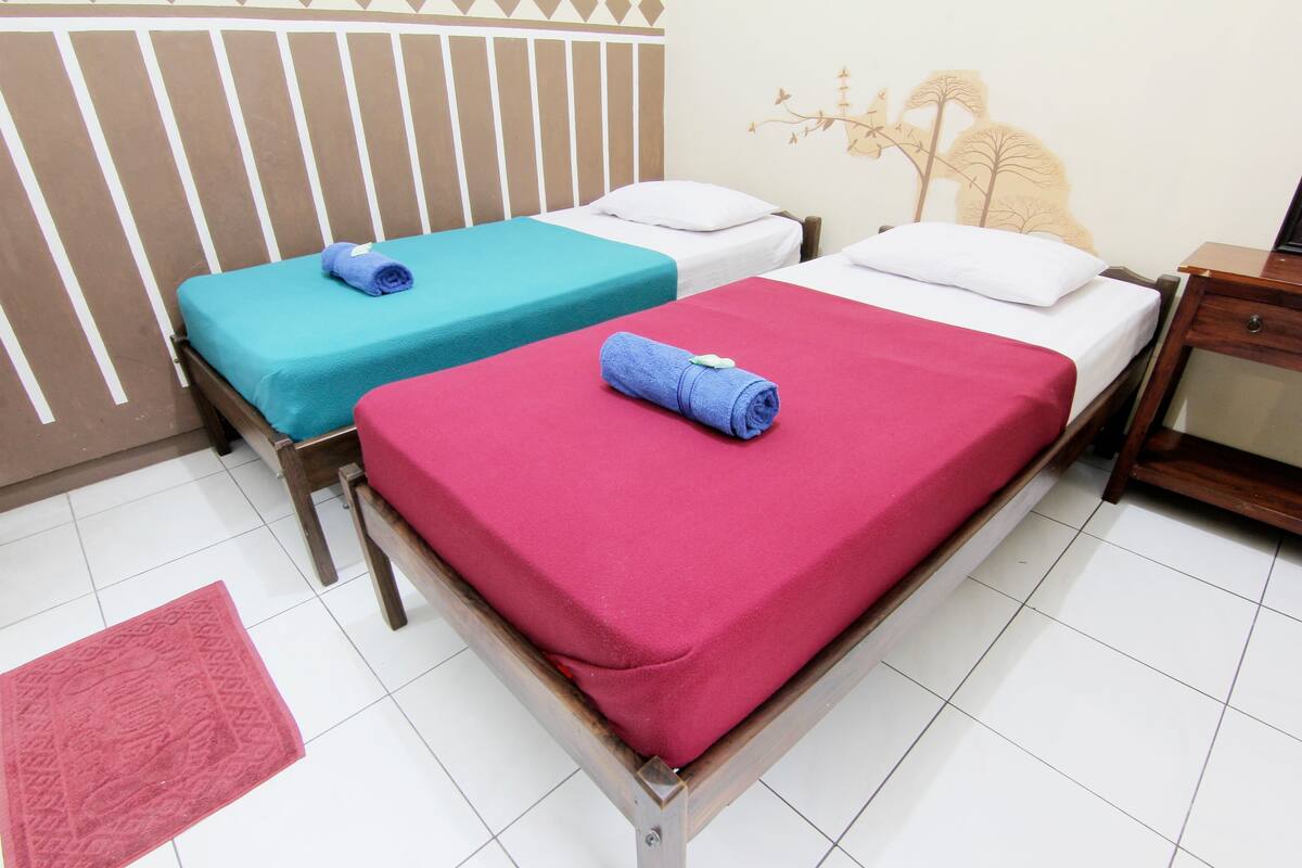 Dos camas individuales con cubrecamas de colores distintos, una mesita de noche de madera y suelo de baldosas.