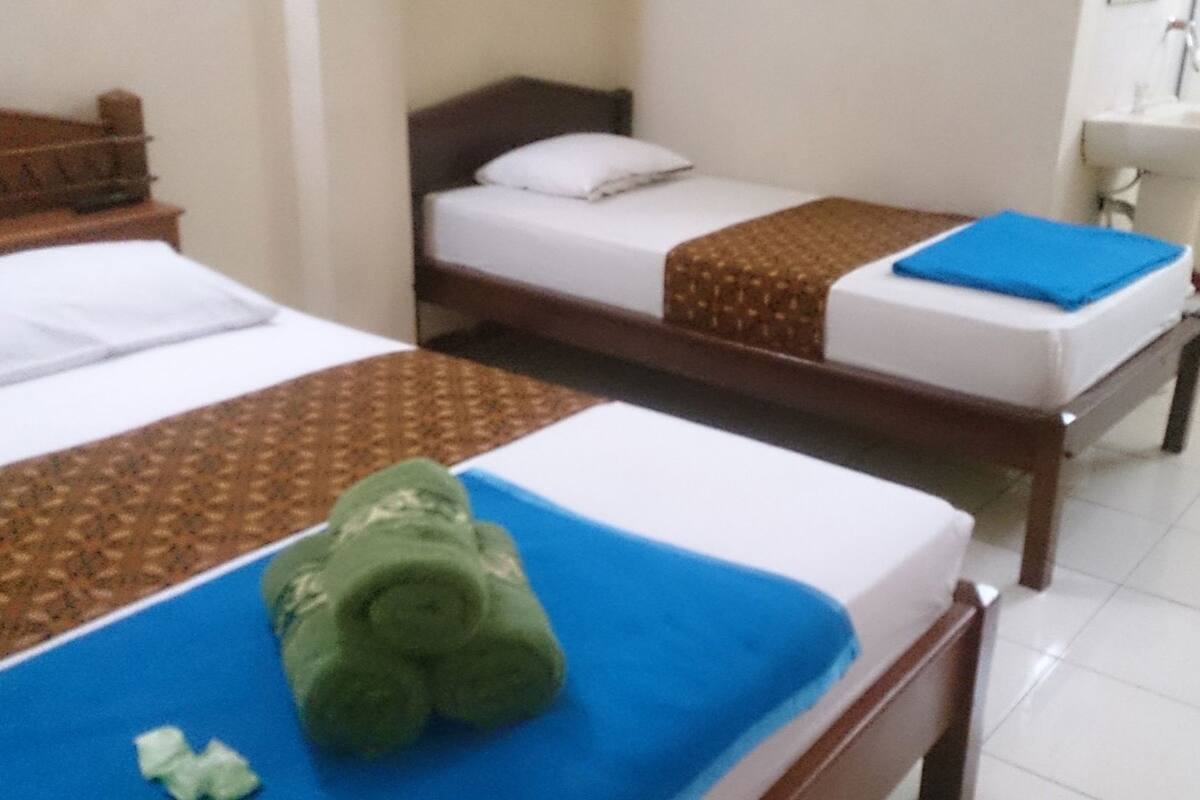 Dos camas individuales con estructuras de madera y ropa de cama blanca, un baño pequeño con lavamanos y una toalla verde sobre una cama.