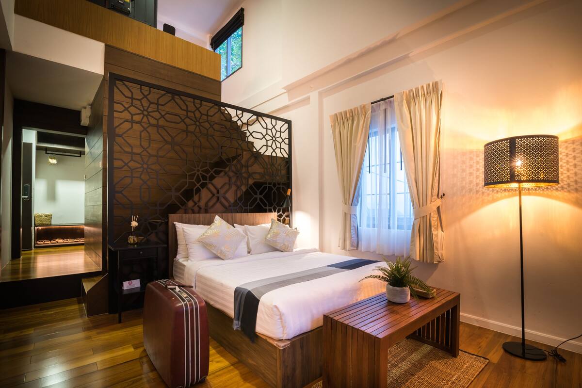 Une chambre d’hôtel moderne avec un lit en bois, un banc, une valise et une lampe sur pied.