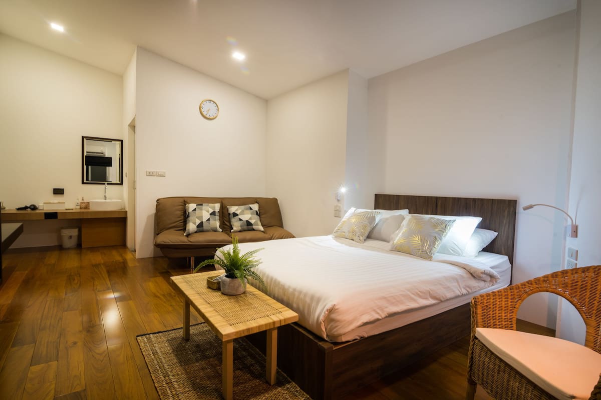 Une chambre moderne avec un plancher en bois, un grand lit avec une literie blanche, une table de chevet en bois, une chaise en osier et une petite table avec une plante.