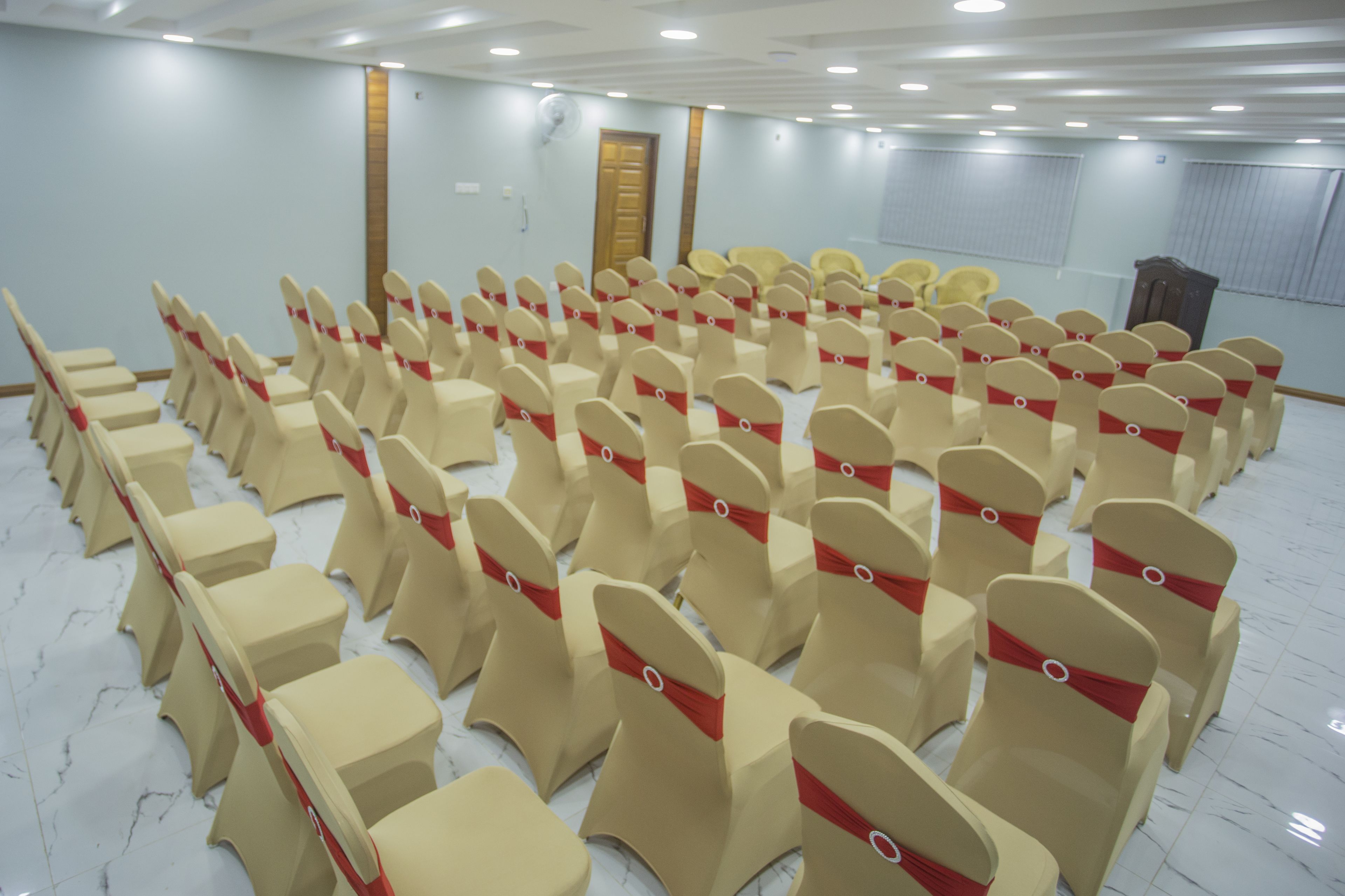 A conference room with rows of beige chairs with red sashes, a wooden door, and a white marble floor.