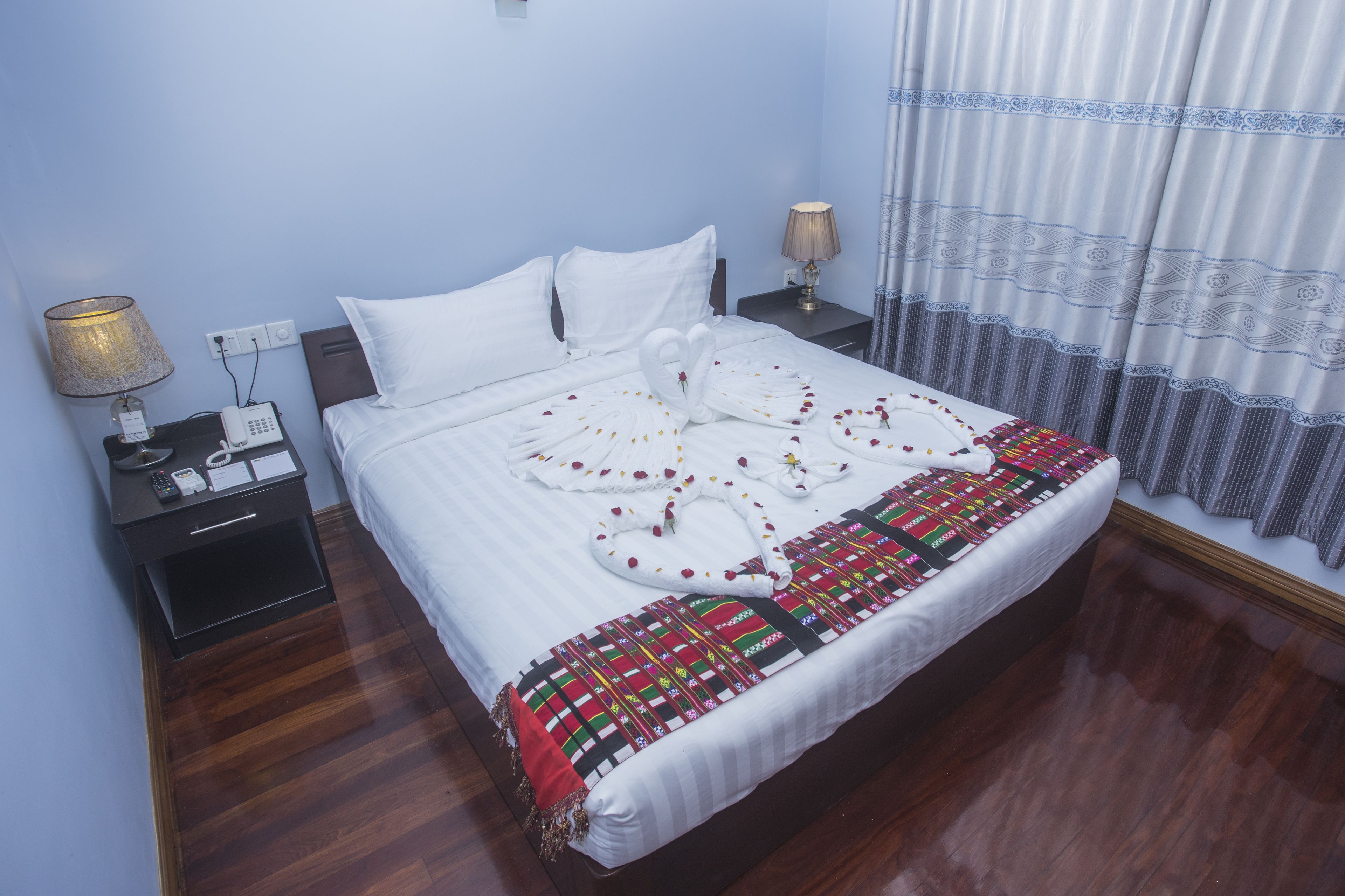 A neatly made bed with white linens and a decorative swan design on the bedspread. There are two bedside tables with lamps and a phone. The room has wooden flooring and light blue walls.