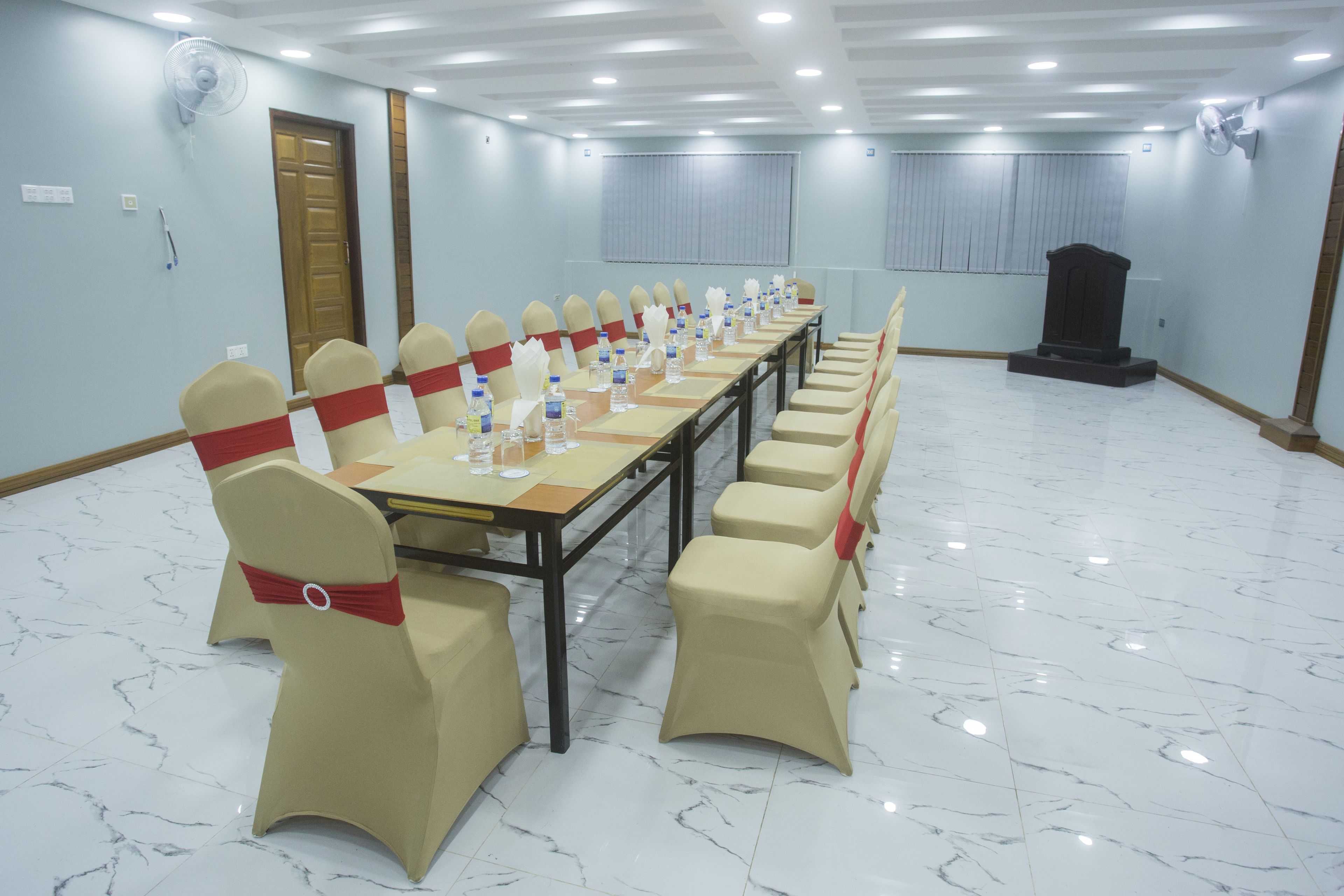 A conference room with a long table, beige chairs with red sashes, and a podium.