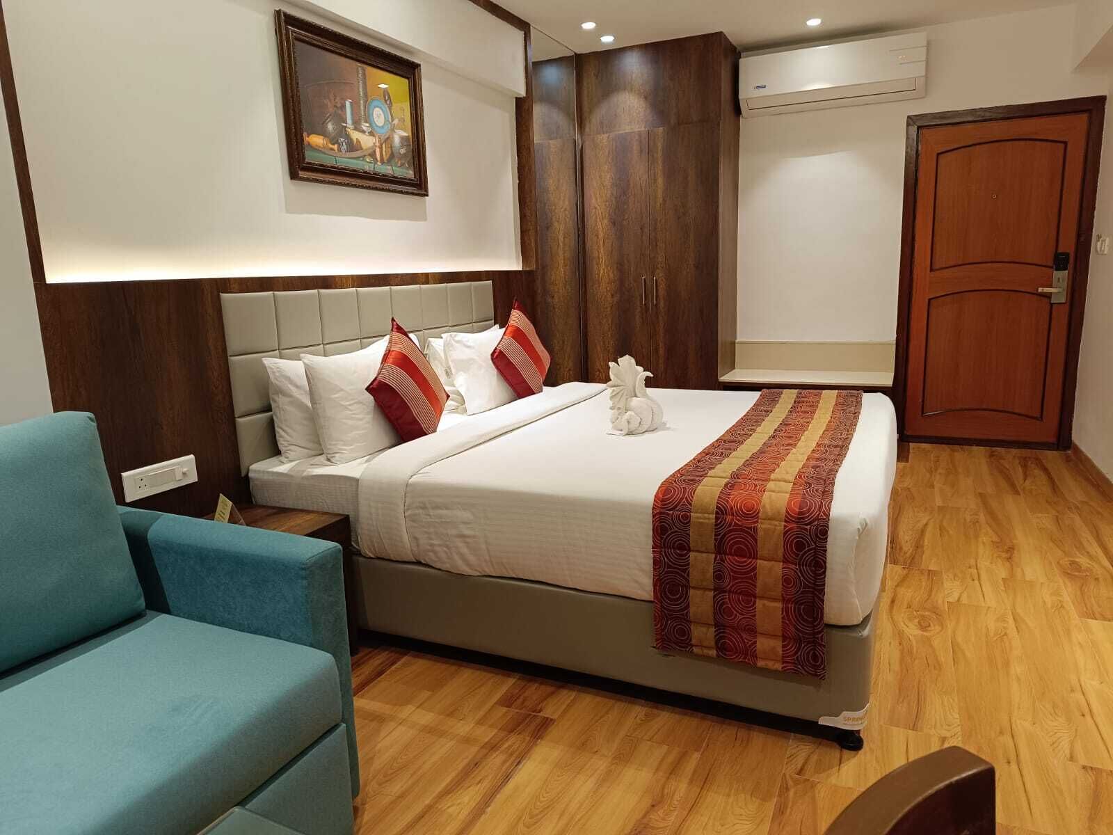Quarto de hotel moderno com uma cama grande, uma mesa de cabeceira, uma poltrona azul, um guarda-roupa de madeira e uma porta.